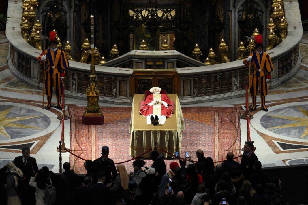 Bento XVI deitado sobre um catafalco, vestido de branco com uma casula vermelha, cor do luto papal, com uma mitra branca debruada a ouro e um rosário entrelaçado nas mãos -